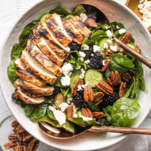 Large salad with spinach, blackberries, cucumber, and grilled chicken, in a white bowl ready to serve.