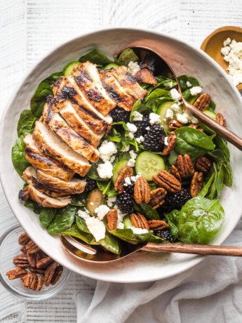 Large salad with spinach, blackberries, cucumber, and grilled chicken, in a white bowl ready to serve.