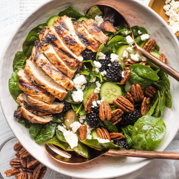 Large salad with spinach, blackberries, cucumber, and grilled chicken, in a white bowl ready to serve.