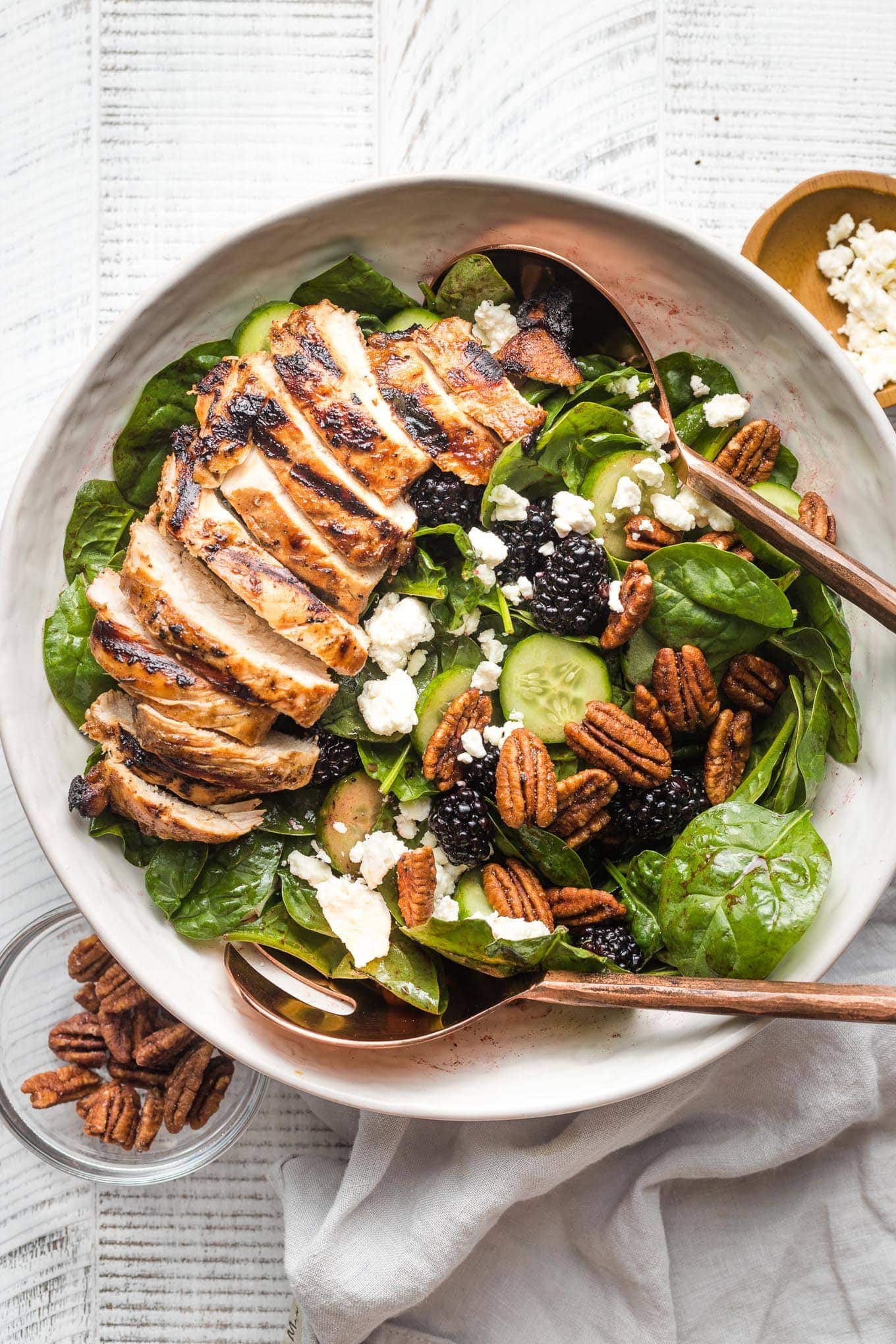 Large salad with spinach, blackberries, cucumber, and grilled chicken, in a white bowl ready to serve.