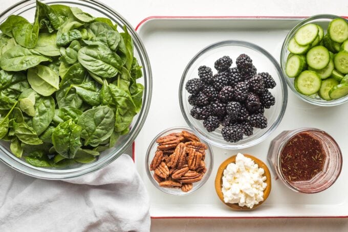 Prep bowls filled with baby spinach, washed blackberries, sliced cucumbers, pecans, crumbled feta, and a jar of dressing.