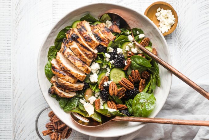 Large salad with spinach, blackberries, cucumber, and grilled chicken, in a white bowl ready to serve.