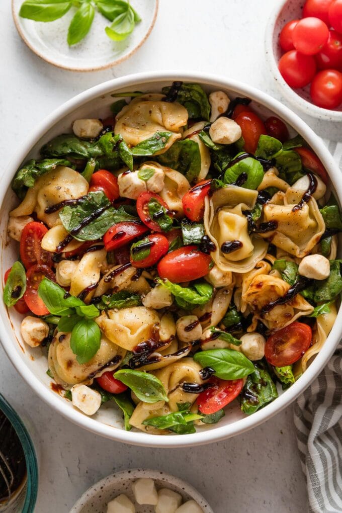 Bowl full of balsamic tortellini Caprese salad, with extra tomatoes, mozzarella, and fresh basil in bowls on the counter nearby.