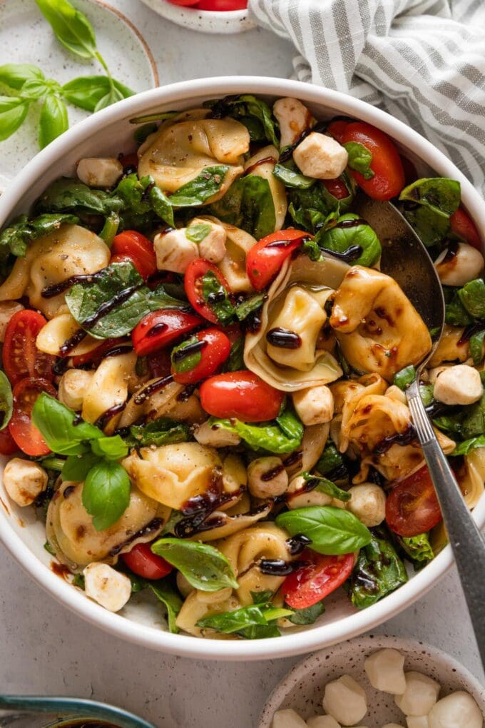 Bowl full of a tortellini Caprese salad with cherry tomatoes, small mozzarella pearls, baby spinach, and a quick homemade balsamic dressing.