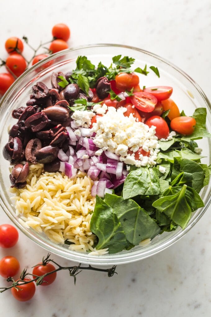Prep bowl containing cooked orzo, halved cherry tomatoes, Kalamata olives, chopped baby spinach, crumbled feta, and a bit of diced red onion.