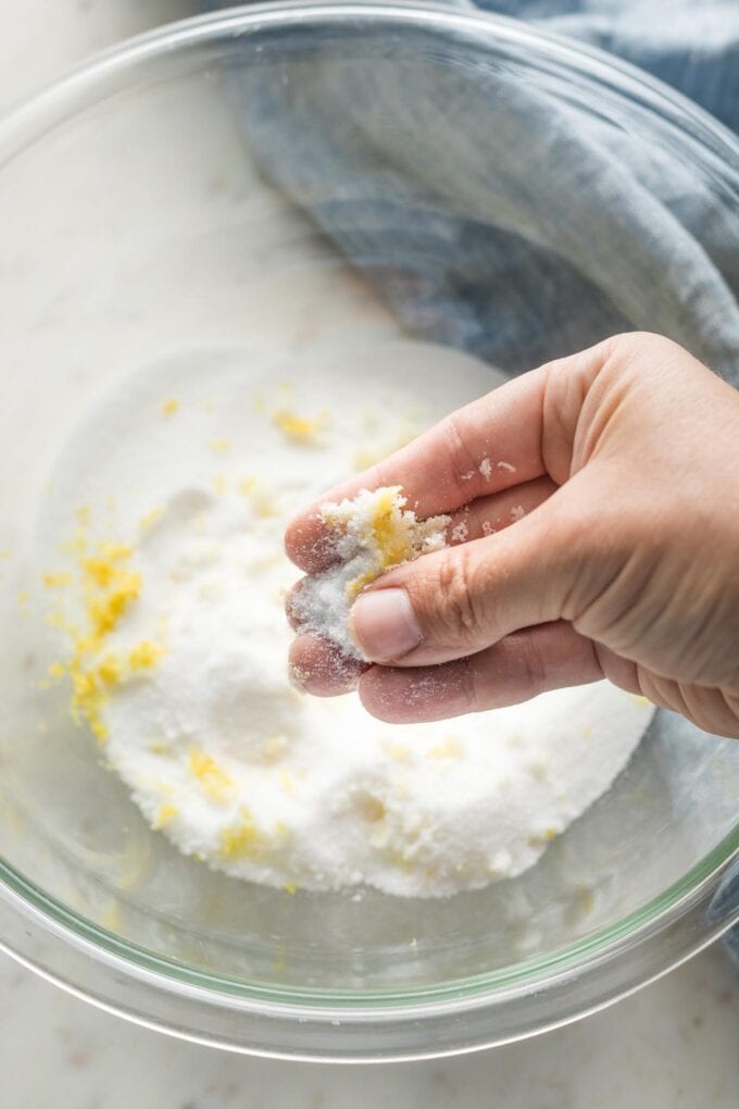 A hand rubbing sugar and lemon zest between the fingers and thumb to infuse the sugar with lemon flavor.