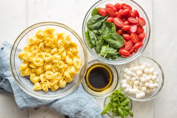 Prep bowls holding cheese tortellini, cherry tomatoes, baby spinach, mozzarella, fresh basil, and ingredients for a simple balsamic dressing.
