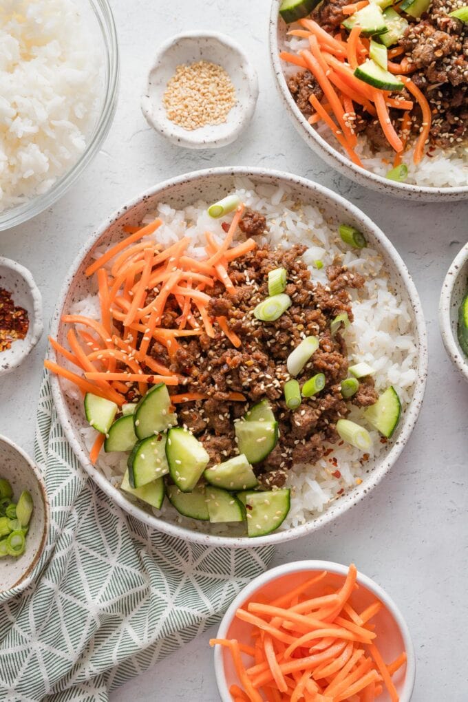 Countertop set with two bowls of Korean-inspired ground beef and rice topped with fresh veggies and toasted sesame seeds.