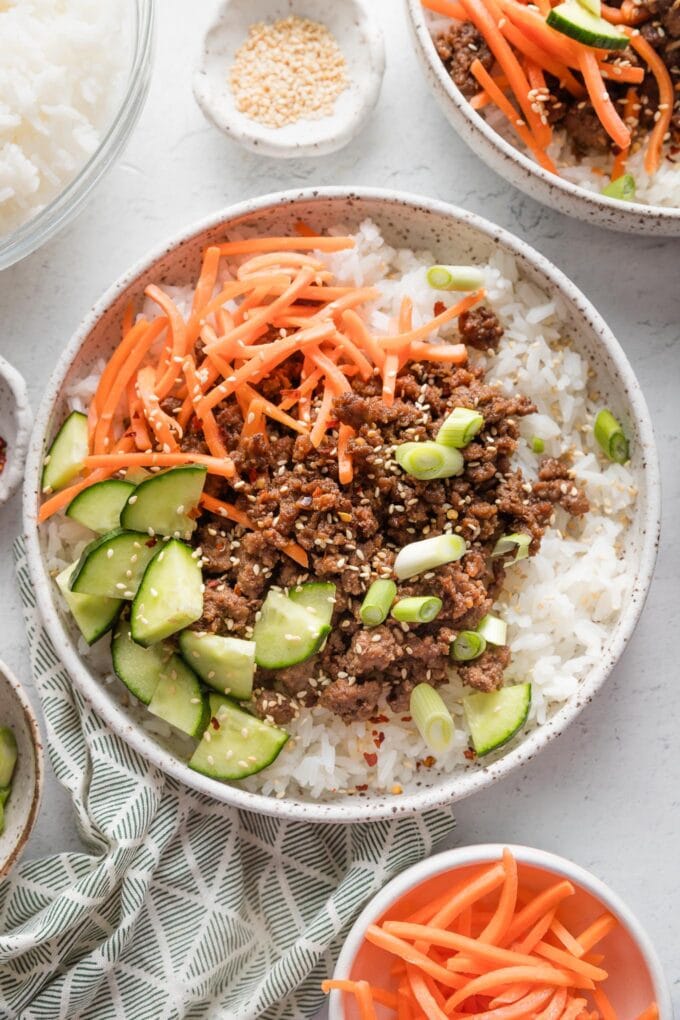 Ceramic bowl full of Korean-inspired ground beef and rice served with cucumber, shredded carrot, and toasted sesame seeds.