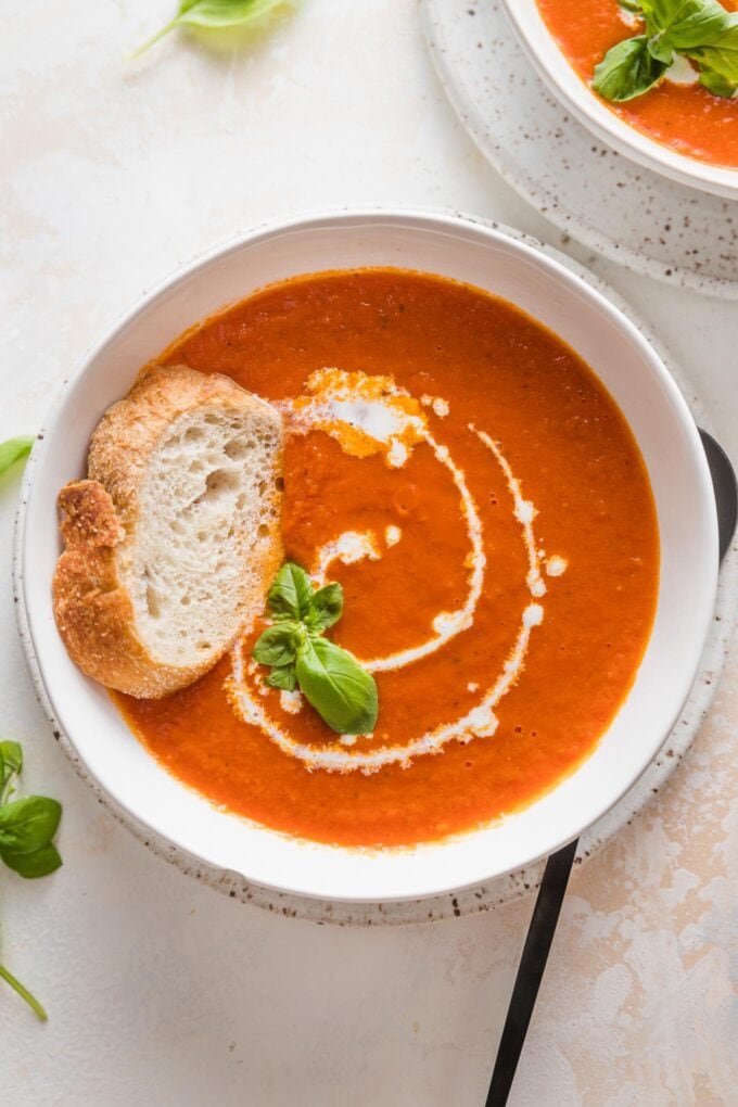 Bowl of roasted tomato basil soup served with fresh herbs, a drizzle of cream, and a slice of baguette.