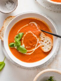Bowl of roasted tomato basil soup served with fresh herbs, a drizzle of cream, and a slice of baguette.