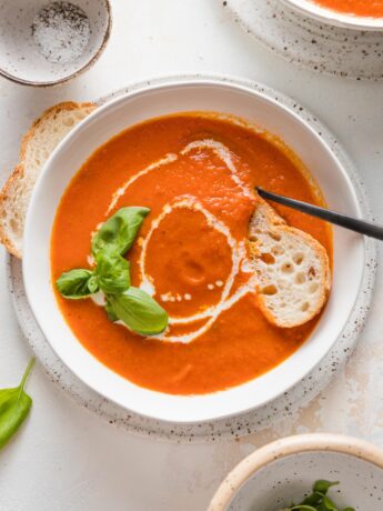 Bowl of roasted tomato basil soup served with fresh herbs, a drizzle of cream, and a slice of baguette.
