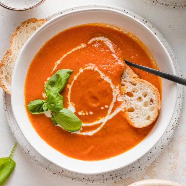 Bowl of roasted tomato basil soup served with fresh herbs, a drizzle of cream, and a slice of baguette.