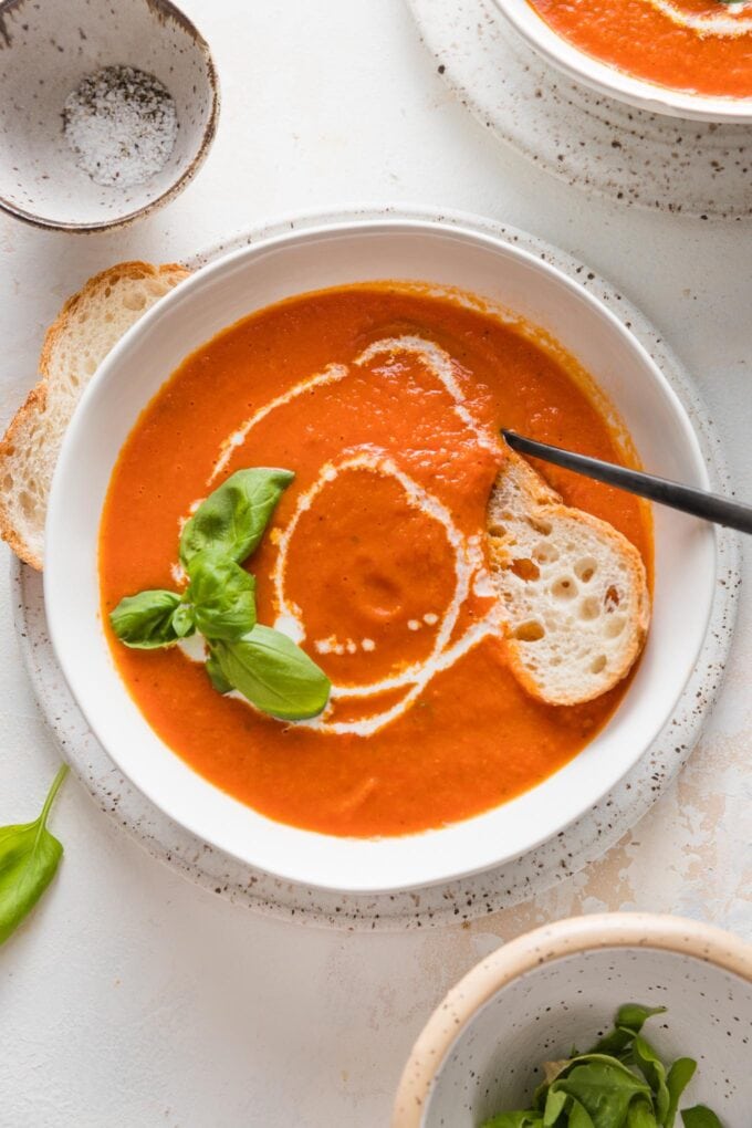 Bowl of roasted tomato basil soup served with fresh herbs, a drizzle of cream, and a slice of baguette.