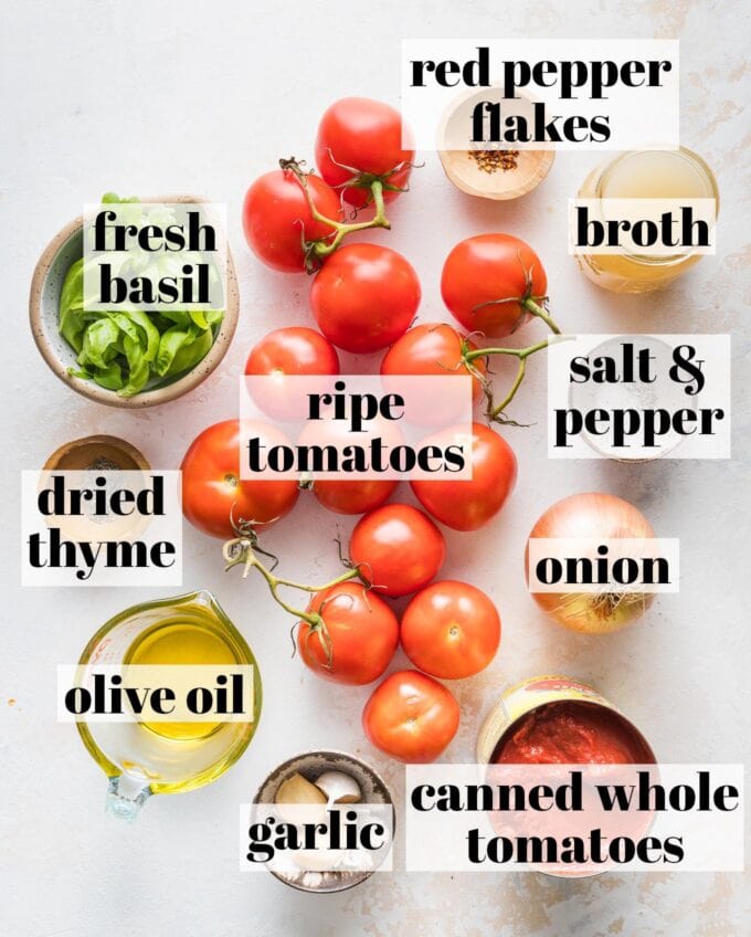 Overhead labeled image of ripe on the vine tomatoes, fresh basil, dried thyme, red pepper flakes, broth, olive oil, a yellow onion, garlic cloves, and canned whole tomatoes in prep bowls and ready to chop and cook.