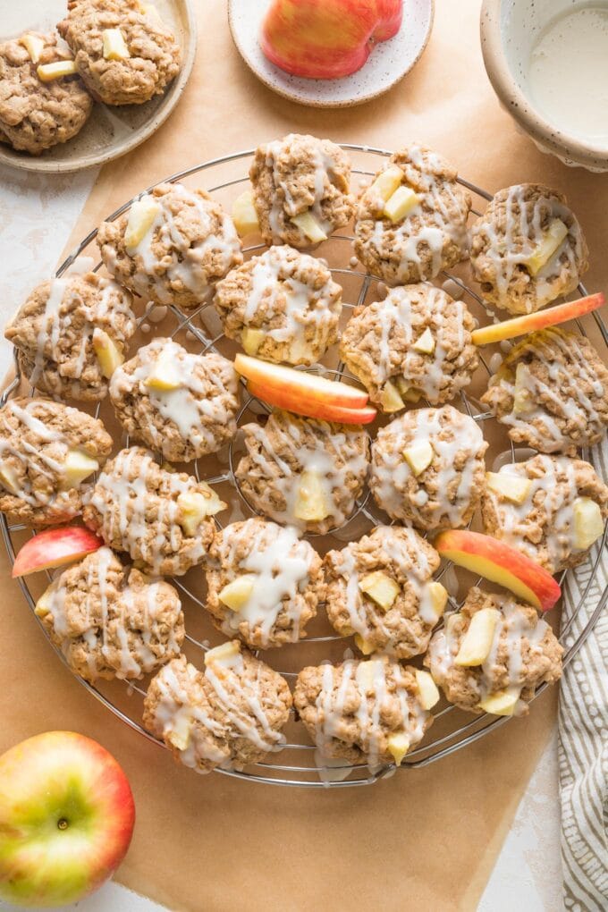 Cooling rack filled with apple cinnamon cookies drizzled with a maple glaze, with apple slices and cinnamon sticks in the background.