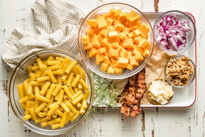 Prep bowls holding dried pasta, cubed butternut squash, chopped red onion, rosemary, bacon, walnuts, and Parmesan cheese.