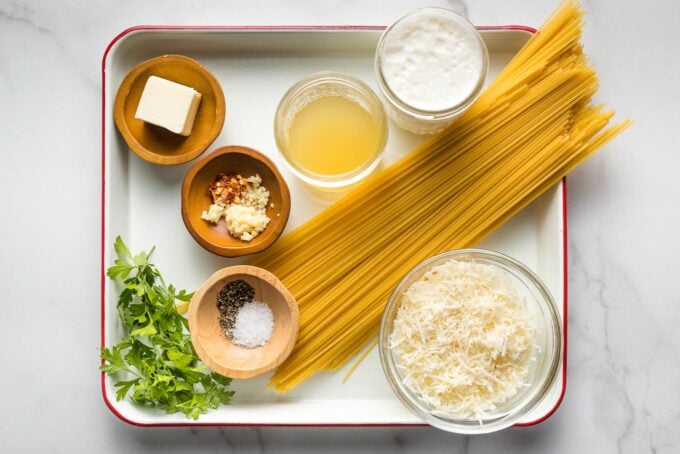 Spaghetti, grated Parmesan cheese, fresh parsley, chicken broth, cream, butter, garlic, and red pepper flakes arranged in prep bowls on a small tray.
