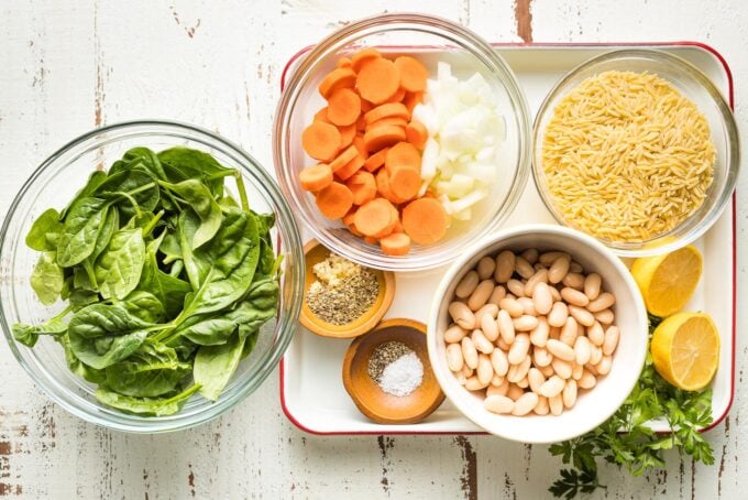 Prep bowls holding baby spinach, chopped carrots and onion, orzo, cannellini beans, and seasonings.