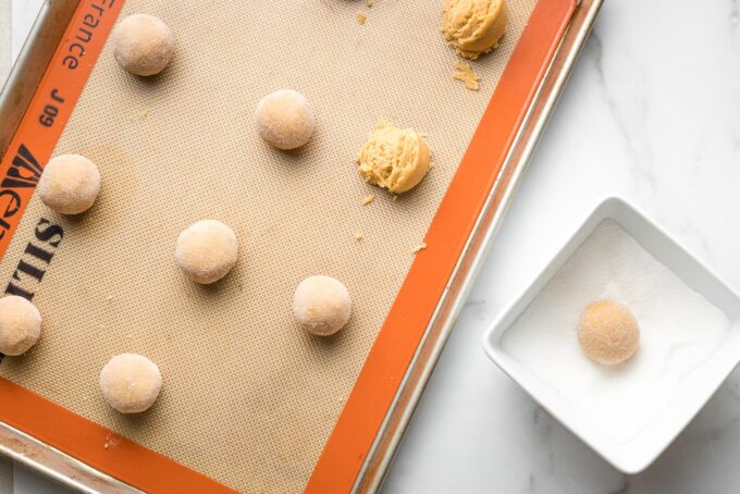 Rolled peanut butter cookies on a baking sheet, with one being dipped in sugar.