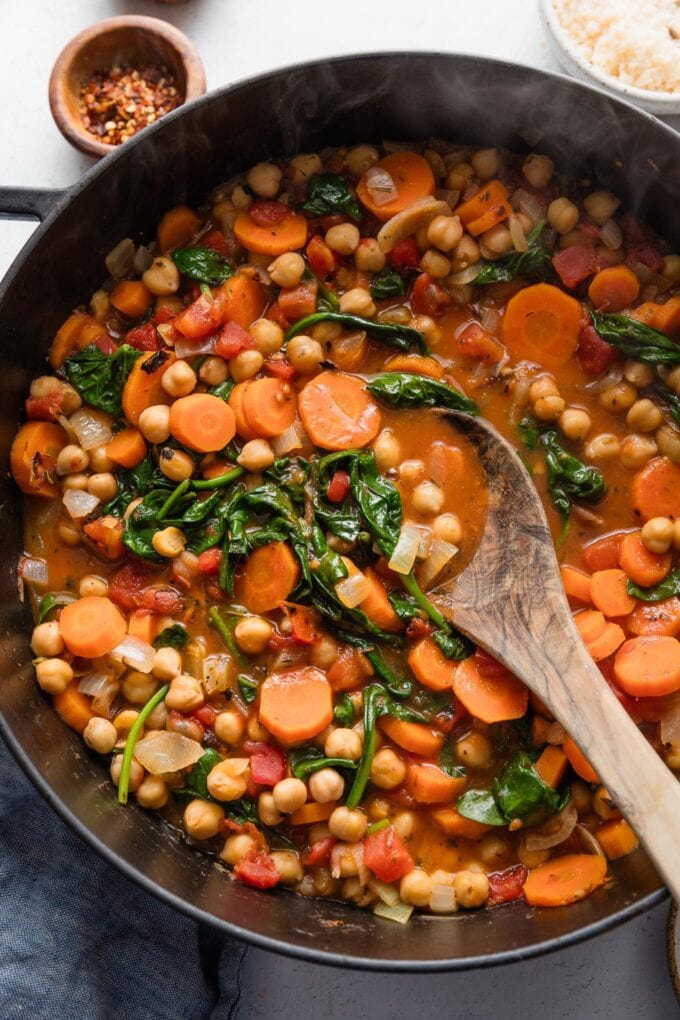 Olive wood spoon stirring a large batch of Tuscan chickpea stew in a deep cast iron skillet.