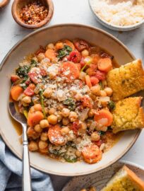 Bowl full of a Tuscan-inspired chickpea stew, with tender carrots, tomatoes, onion, garlic, baby spinach, and Parmesan on top, with garlic bread pieces on the side of the bowl.