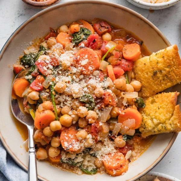 Bowl full of a Tuscan-inspired chickpea stew, with tender carrots, tomatoes, onion, garlic, baby spinach, and Parmesan on top, with garlic bread pieces on the side of the bowl.