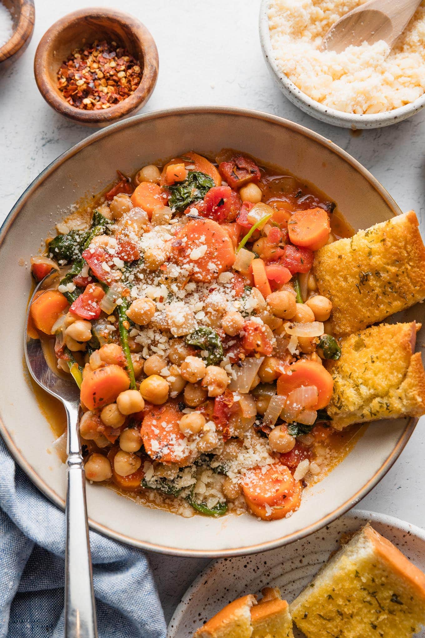 Bowl full of a Tuscan-inspired chickpea stew, with tender carrots, tomatoes, onion, garlic, baby spinach, and Parmesan on top, with garlic bread pieces on the side of the bowl.