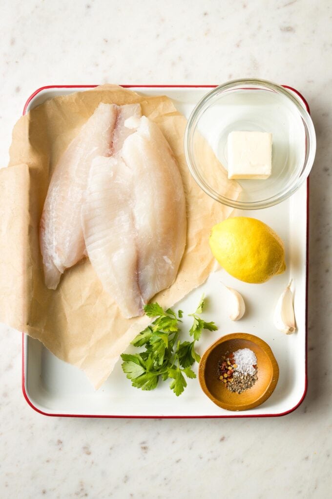 Tilapia filets, butter, lemon, garlic, parsley, and seasonings prepped on a tray.