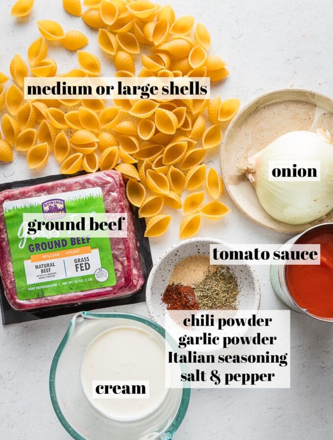 Labeled overhead photo of ground beef, uncooked pasta shells, a half a yellow onion, tomato sauce, a splash of cream, and spices.