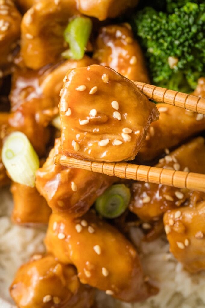 Close-up of a bite-sized piece of chicken coated in teriyaki sauce, held by a pair of chopsticks.