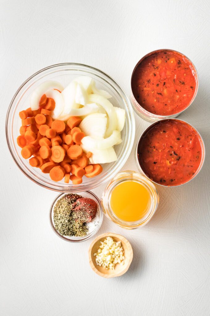 Prep bowls holding chopped carrots, onion, broth, garlic, and seasonings, plus two large cans of fire-roasted tomatoes.