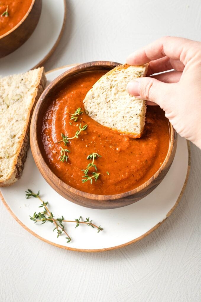 Hand dipping a slice of bread into a bowl of tomato soup.
