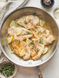 Skillet of lemon rosemary chicken ready to serve, with extra fresh herbs and lemon wedges for garnish.