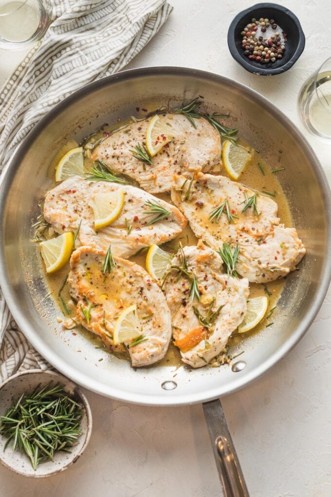 Skillet of lemon rosemary chicken ready to serve, with extra fresh herbs and lemon wedges for garnish.