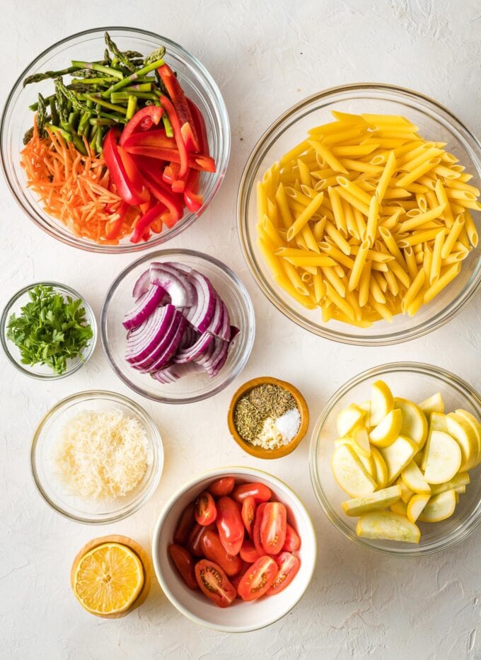 Prep bowls containing dried penne, carrots, asparagus, red bell pepper, yellow squash, red onion, lemon, garlic, and herbs.