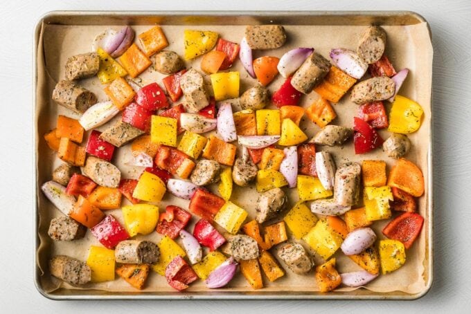 Sausages and veggies, uncooked, spread out on a sheet pan.