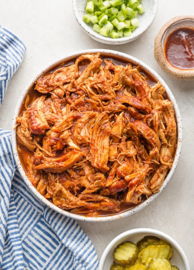 White bowl full of BBQ pulled chicken made in a slow cooker, with green onions, pickles, and extra BBQ sauce nearby for garnish.