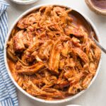 White bowl full of BBQ pulled chicken made in a slow cooker, with green onions, pickles, and extra BBQ sauce nearby for garnish.