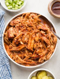 White bowl full of BBQ pulled chicken made in a slow cooker, with green onions, pickles, and extra BBQ sauce nearby for garnish.