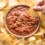 A chip being dipped into a bowl of restaurant-style blender salsa, surrounded by chips.