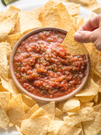 A chip being dipped into a bowl of restaurant-style blender salsa, surrounded by chips.