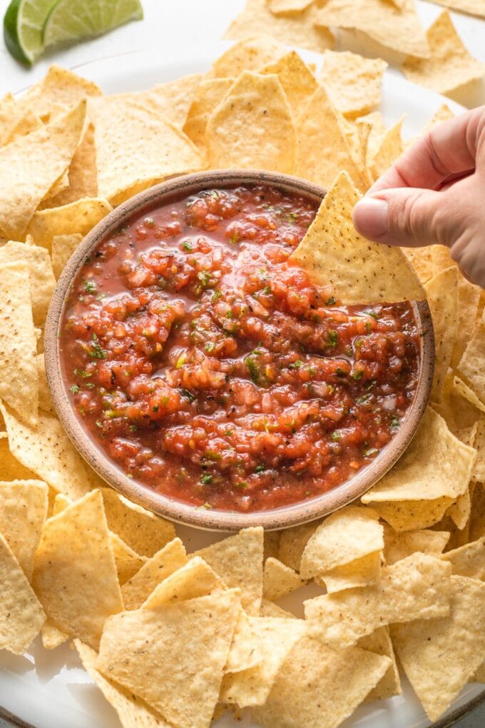 A chip being dipped into a bowl of restaurant-style blender salsa, surrounded by chips.