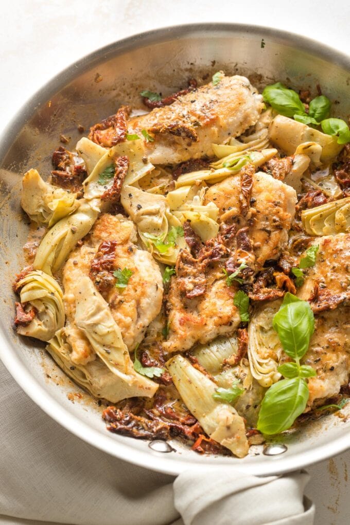 Skillet with cooked chicken breasts, sun-dried tomatoes, and artichokes in a light cream sauce, garnished with basil.