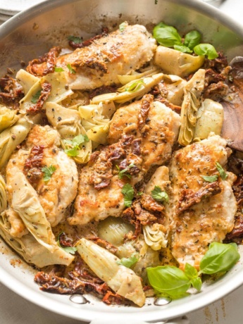 Skillet with cooked chicken breasts, sun-dried tomatoes, and artichokes in a light cream sauce, garnished with basil.