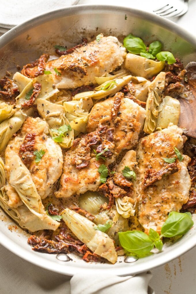 Skillet with cooked chicken breasts, sun-dried tomatoes, and artichokes in a light cream sauce, garnished with basil.