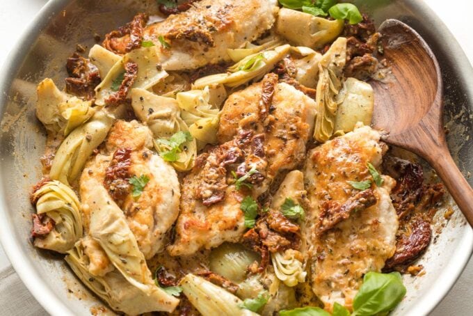 Close-up of finished chicken with sun-dried tomatoes and artichokes in skillet, with a spoon ready to lift up a piece and serve.