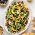 White platter with a peach arugula salad, with prosciutto, blueberries, pine nuts, and a white wine vinaigrette, ready to serve.