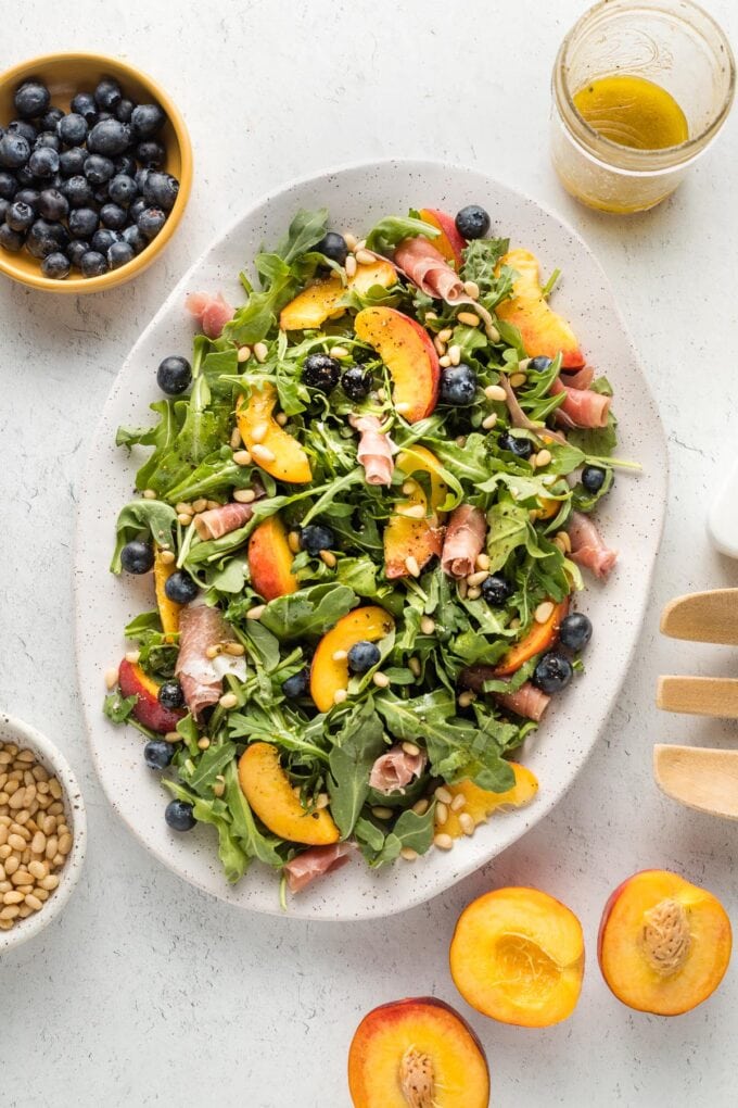 White platter with a peach arugula salad, with prosciutto, blueberries, pine nuts, and a white wine vinaigrette, ready to serve.