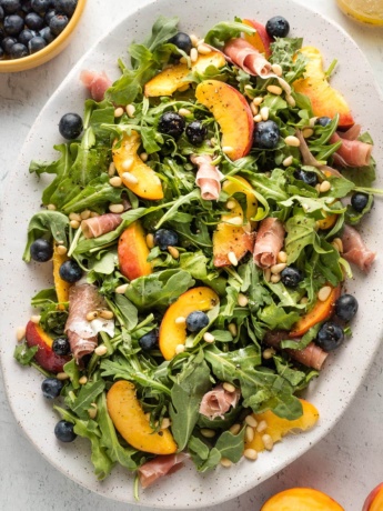 White platter with a peach arugula salad, with prosciutto, blueberries, pine nuts, and a white wine vinaigrette, ready to serve.